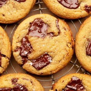 Close-up overhead view of a golden brown chocolate chunk cookie on a cooling rack, topped with glossy melted chocolate and a sprinkle of flaky sea salt.