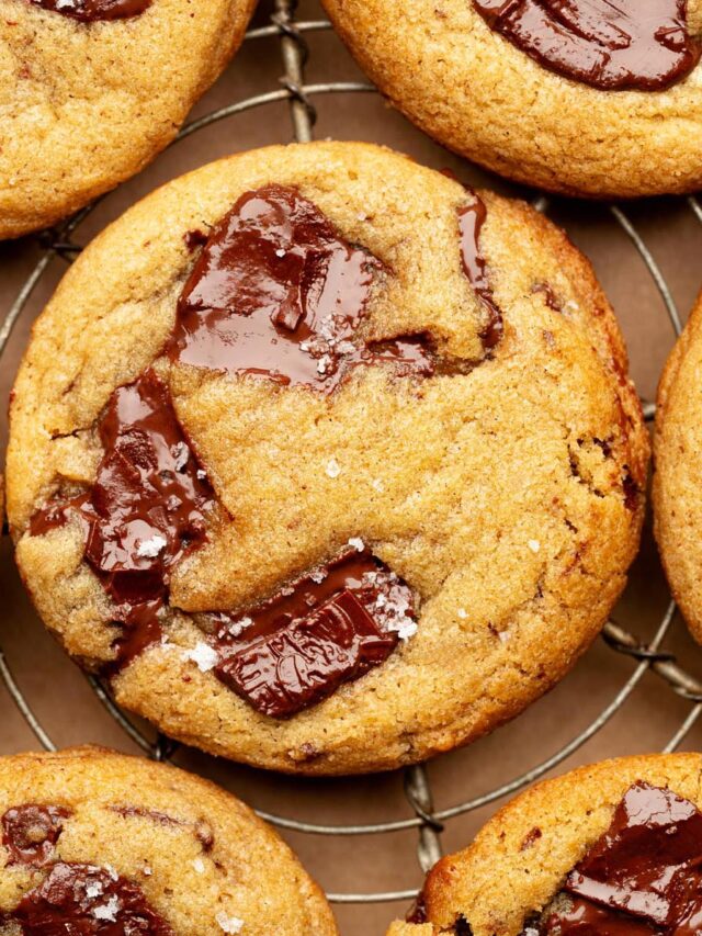 Close-up overhead view of a golden brown chocolate chunk cookie on a cooling rack, topped with glossy melted chocolate and a sprinkle of flaky sea salt.