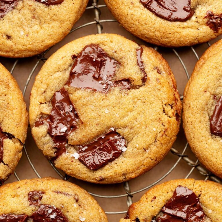 Close-up overhead view of a golden brown chocolate chunk cookie on a cooling rack, topped with glossy melted chocolate and a sprinkle of flaky sea salt.