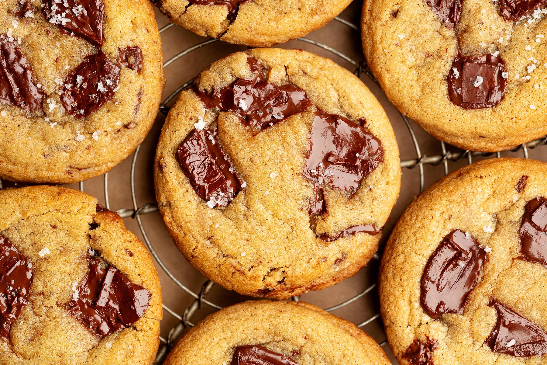 Overhead close-up of freshly baked chocolate chunk cookies on a cooling rack, topped with melted chocolate puddles and a sprinkle of flaky sea salt.