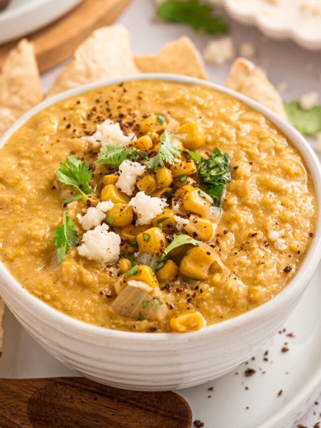 Bowl of creamy Mexican street corn chowder topped with corn, cilantro, and Cotija cheese, served with tortilla chips and a wooden spoon.