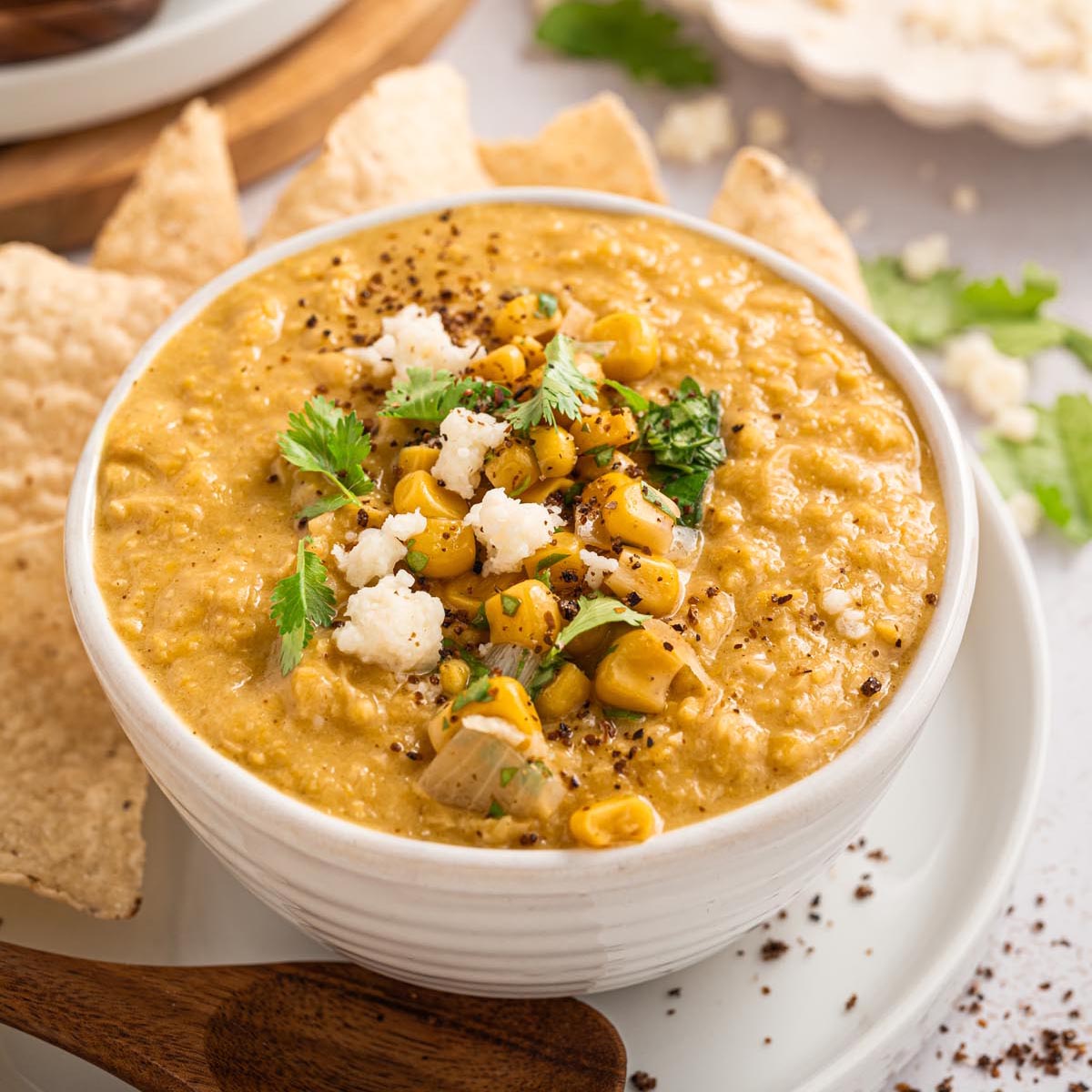 Bowl of creamy Mexican street corn chowder topped with corn, cilantro, and Cotija cheese, served with tortilla chips and a wooden spoon.
