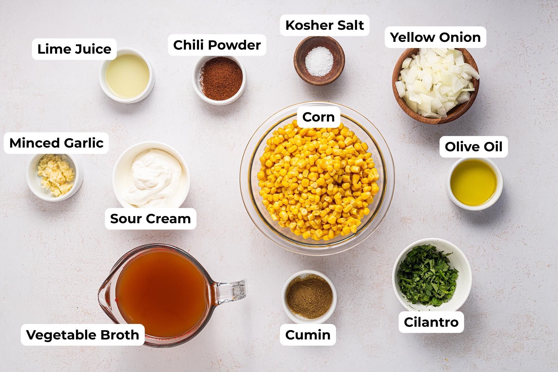 Labeled overhead image of Mexican Street Corn Chowder ingredients, including corn, yellow onion, olive oil, minced garlic, sour cream, vegetable broth, lime juice, cumin, chili powder, kosher salt, and chopped cilantro arranged on a light surface.