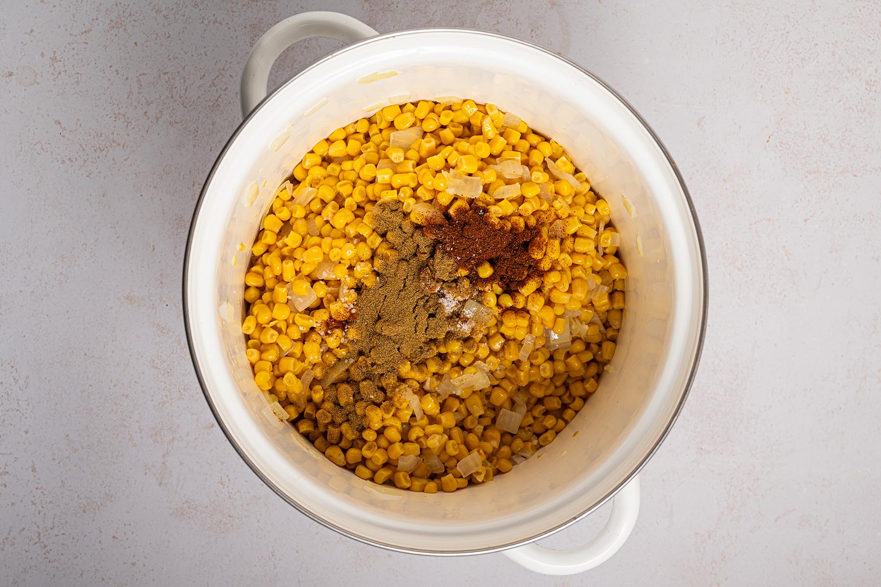 Pot filled with corn and sautéed onions topped with cumin, chili powder, and salt before stirring.