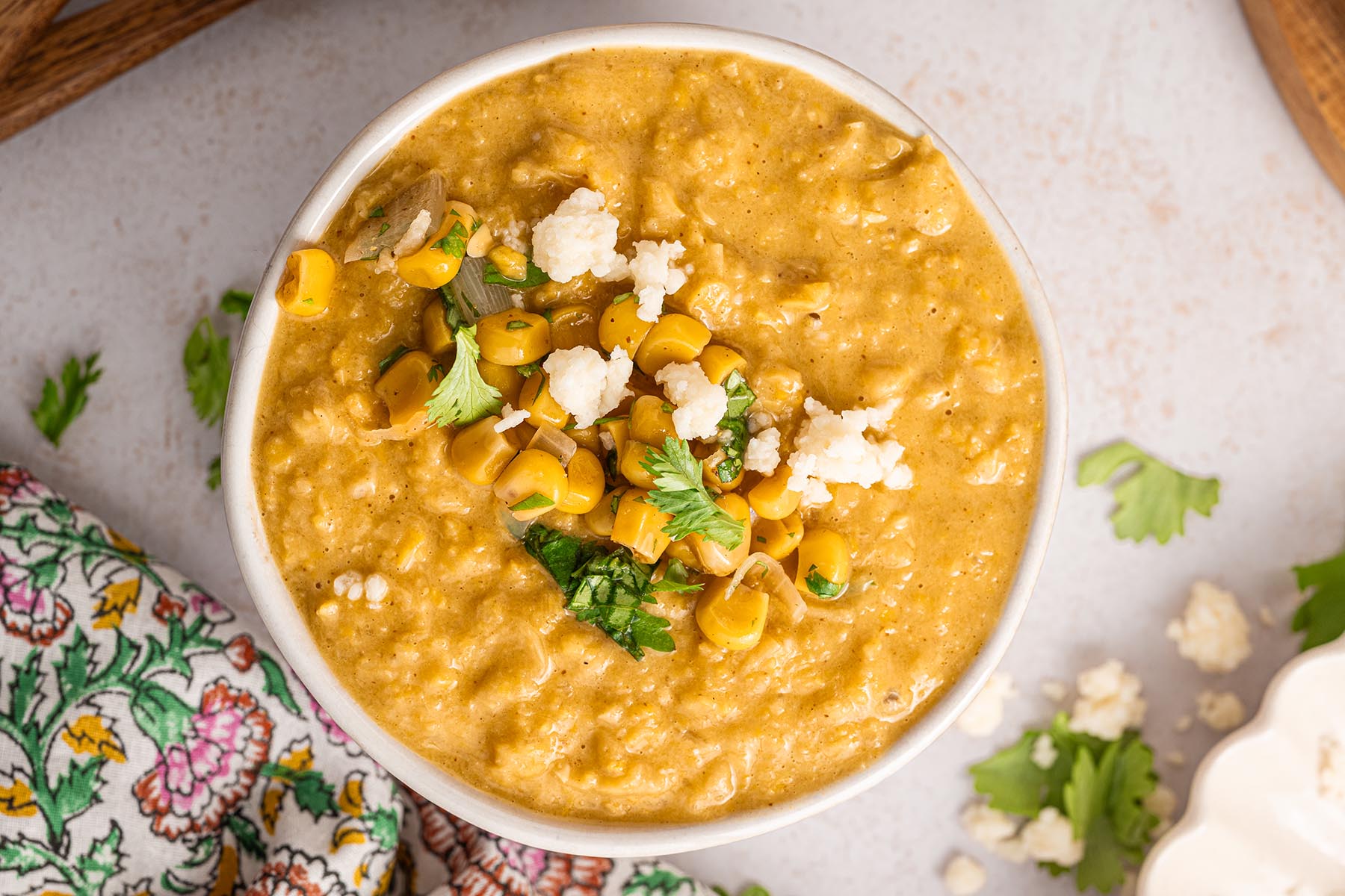 Overhead view of Mexican street corn chowder in a white bowl, garnished with corn, cilantro, and Cotija, with scattered cheese and herbs on the table.