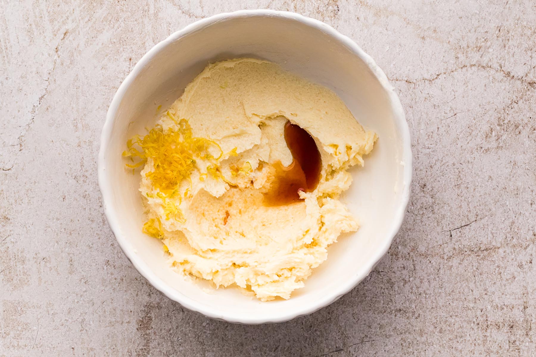 Fresh lemon zest and vanilla extract added to creamed butter and sugar in a white mixing bowl.