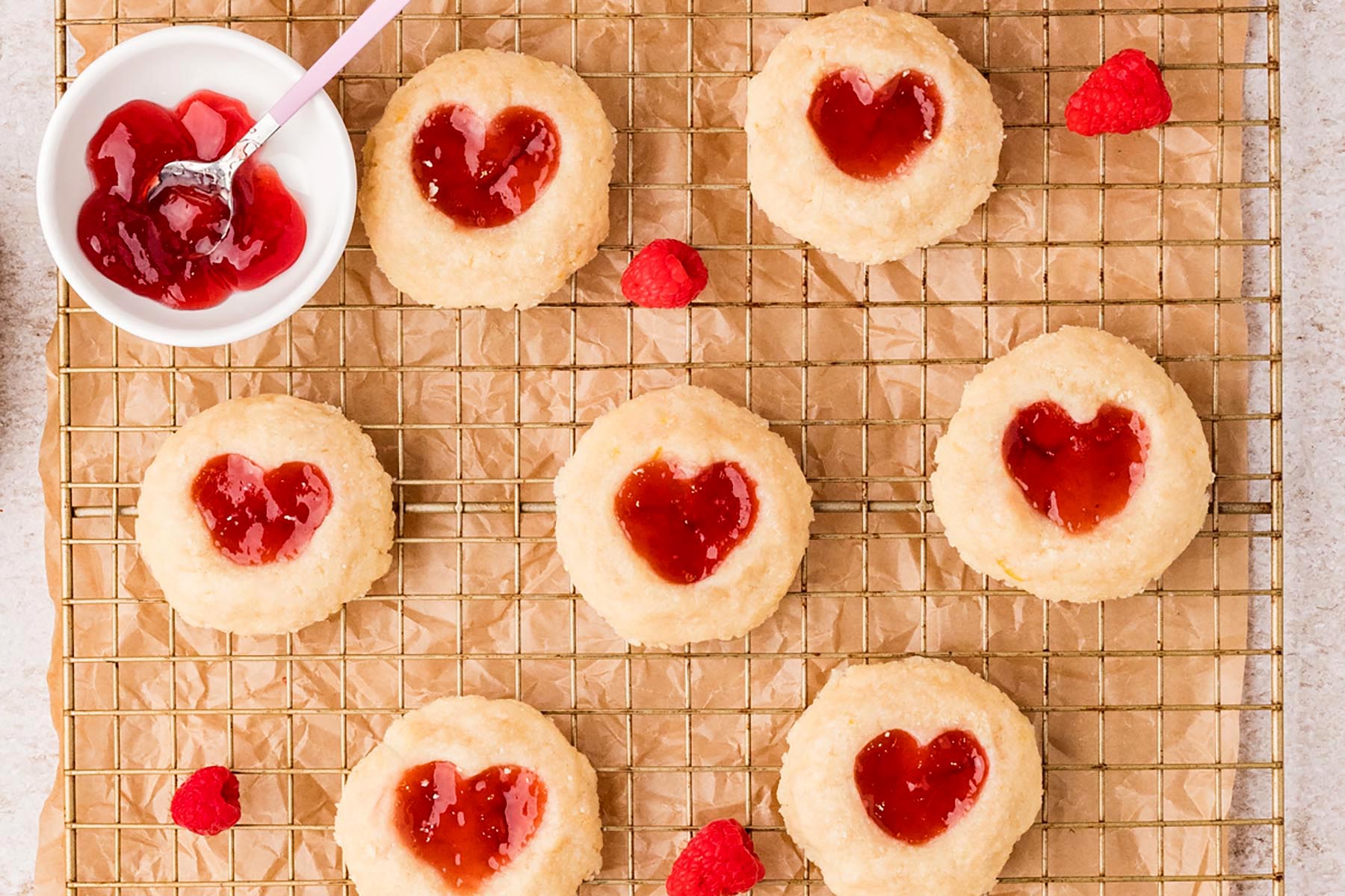 Heart-shaped raspberry thumbprint cookies cooling on a wire rack, filled with glossy raspberry jam.