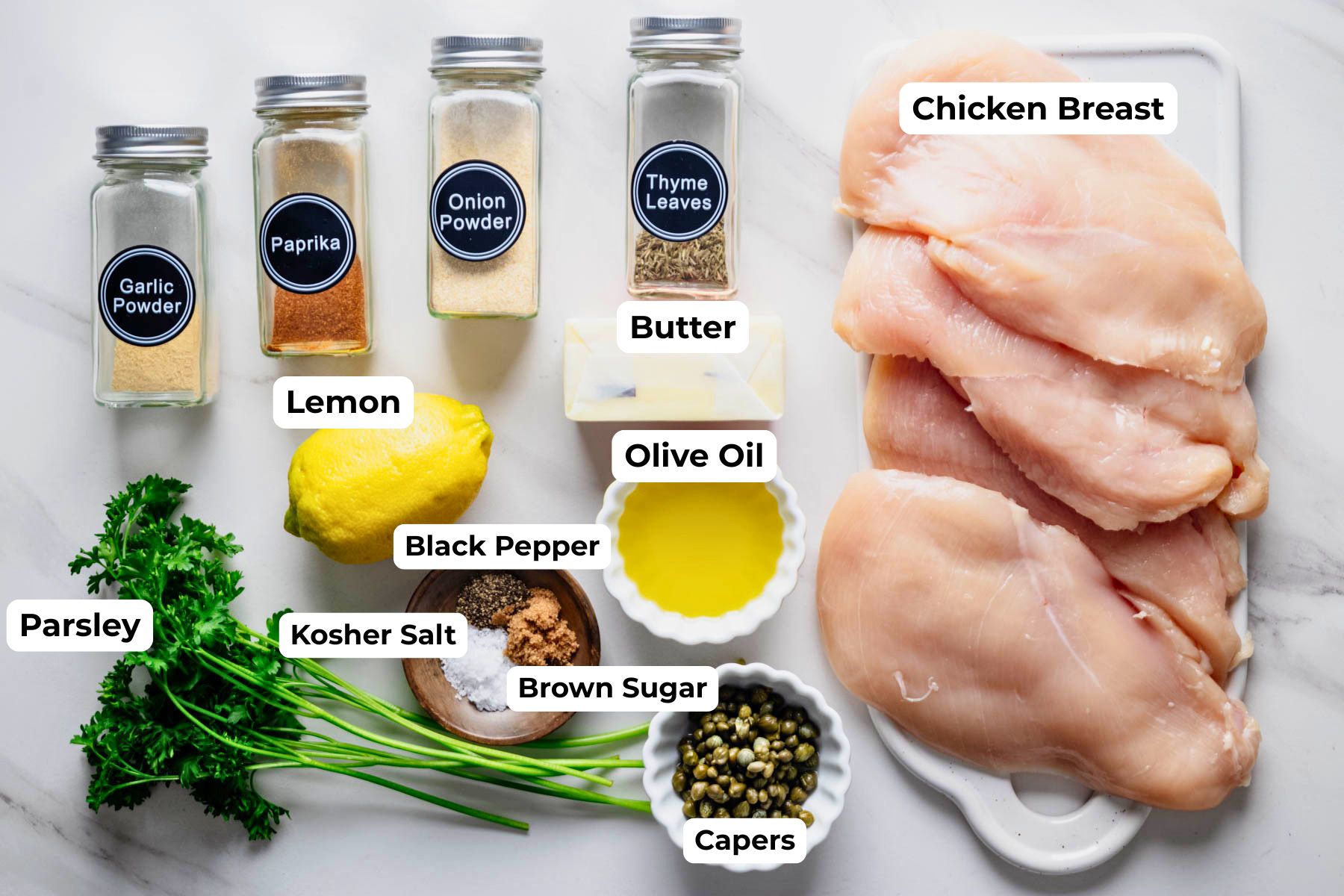 Ingredients for baked chicken piccata arranged on a white surface, including chicken breasts, lemon, capers, parsley, butter, olive oil, and seasonings.