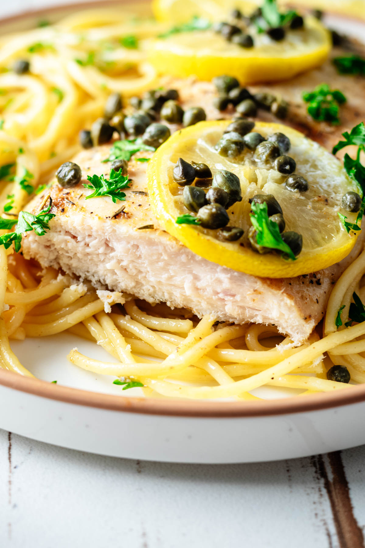 Close-up of sliced baked chicken piccata served over spaghetti with lemon slices, capers, and fresh parsley.