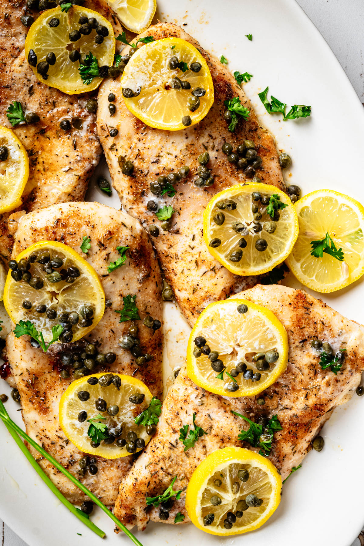 Overhead view of baked chicken piccata topped with lemon slices, capers, and parsley on a white serving platter.