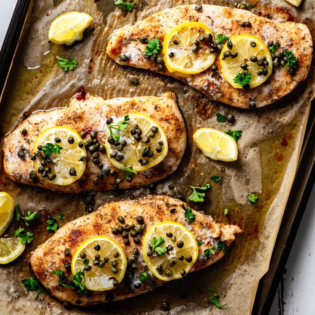 Overhead view of baked chicken piccata on a parchment-lined sheet pan, topped with lemon slices, capers, and fresh parsley with pan juices surrounding the chicken.
