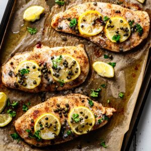 Overhead view of baked chicken piccata on a parchment-lined sheet pan, topped with lemon slices, capers, and fresh parsley with pan juices surrounding the chicken.