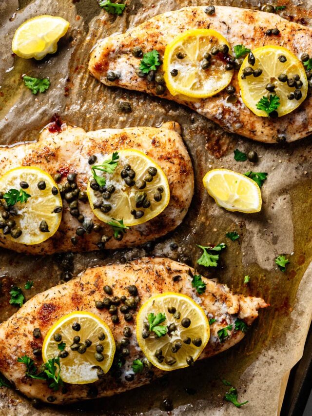 Overhead view of baked chicken piccata on a parchment-lined sheet pan, topped with lemon slices, capers, and fresh parsley with pan juices surrounding the chicken.