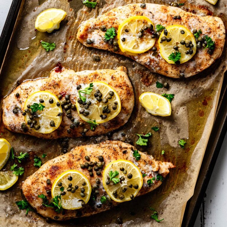 Overhead view of baked chicken piccata on a parchment-lined sheet pan, topped with lemon slices, capers, and fresh parsley with pan juices surrounding the chicken.