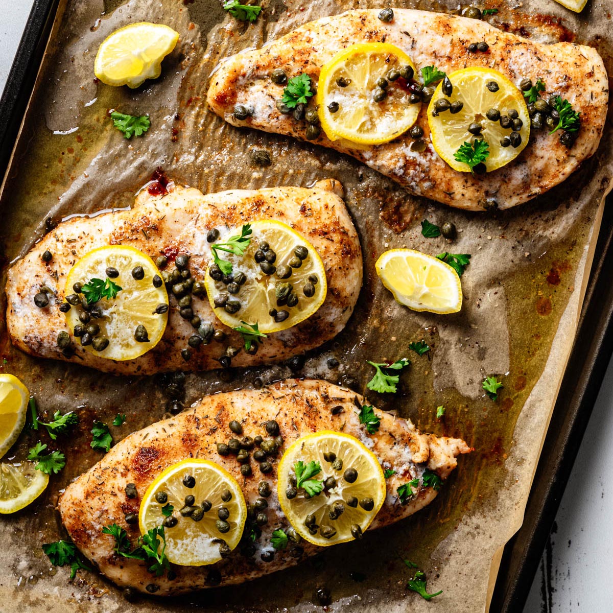 Overhead view of baked chicken piccata on a parchment-lined sheet pan, topped with lemon slices, capers, and fresh parsley with pan juices surrounding the chicken.