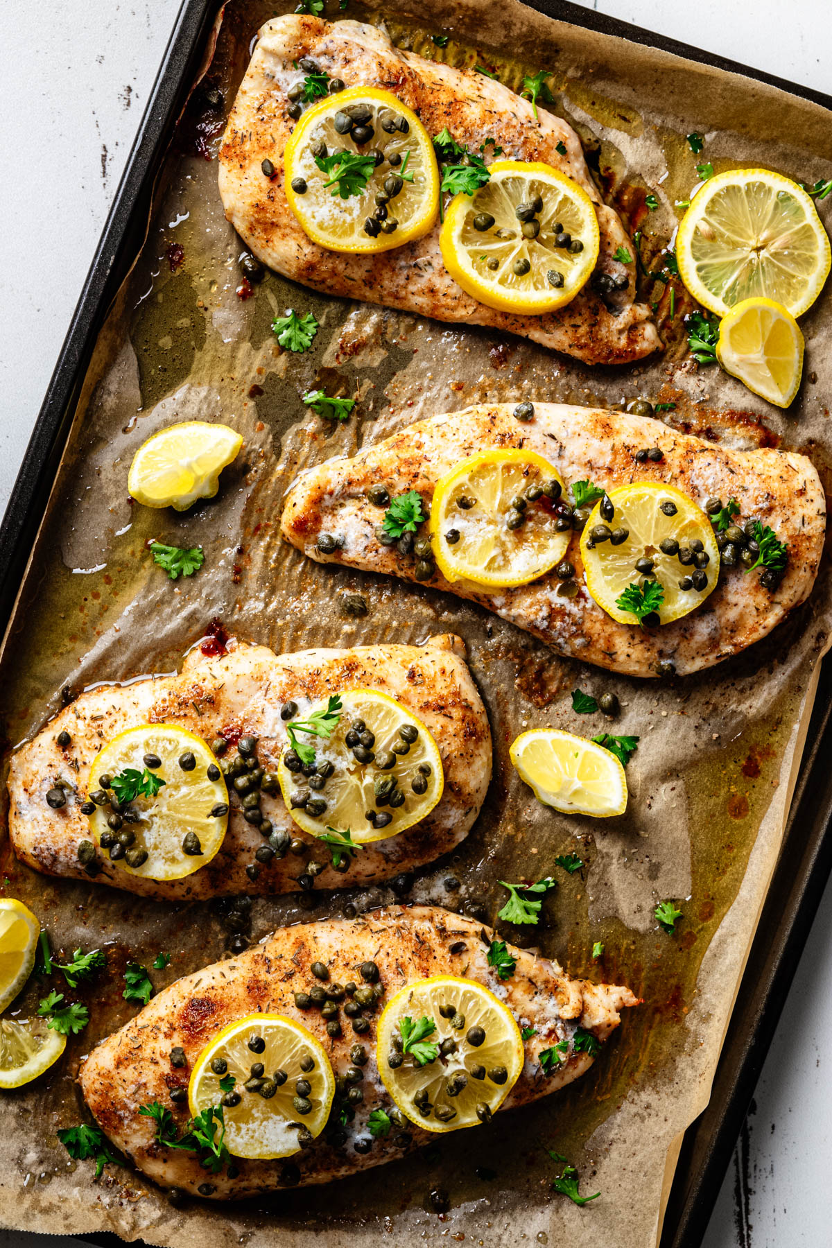 Baked chicken piccata on a parchment-lined sheet pan topped with lemon slices, capers, and fresh parsley.