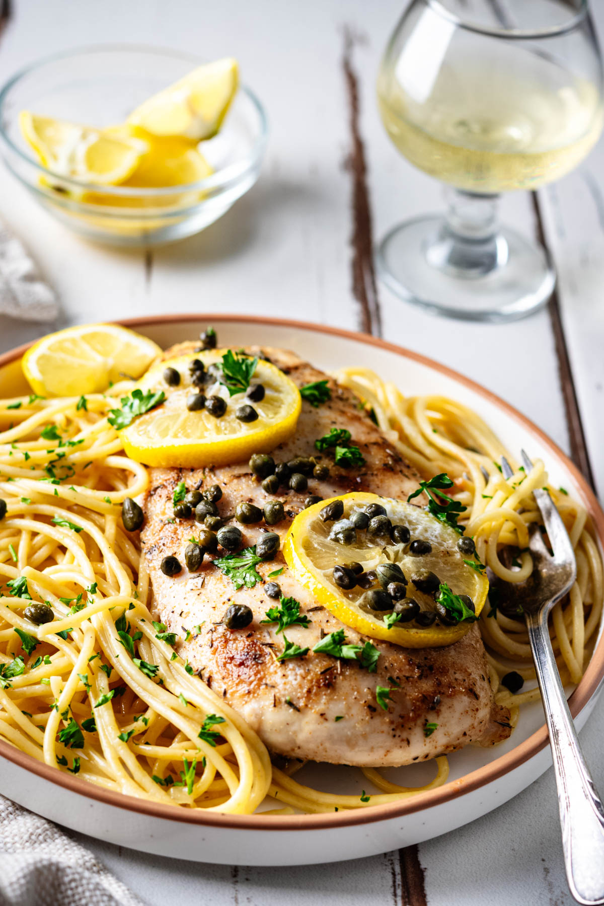 Plated baked chicken piccata served over buttered spaghetti with lemon slices, capers, parsley, and a glass of white wine in the background.