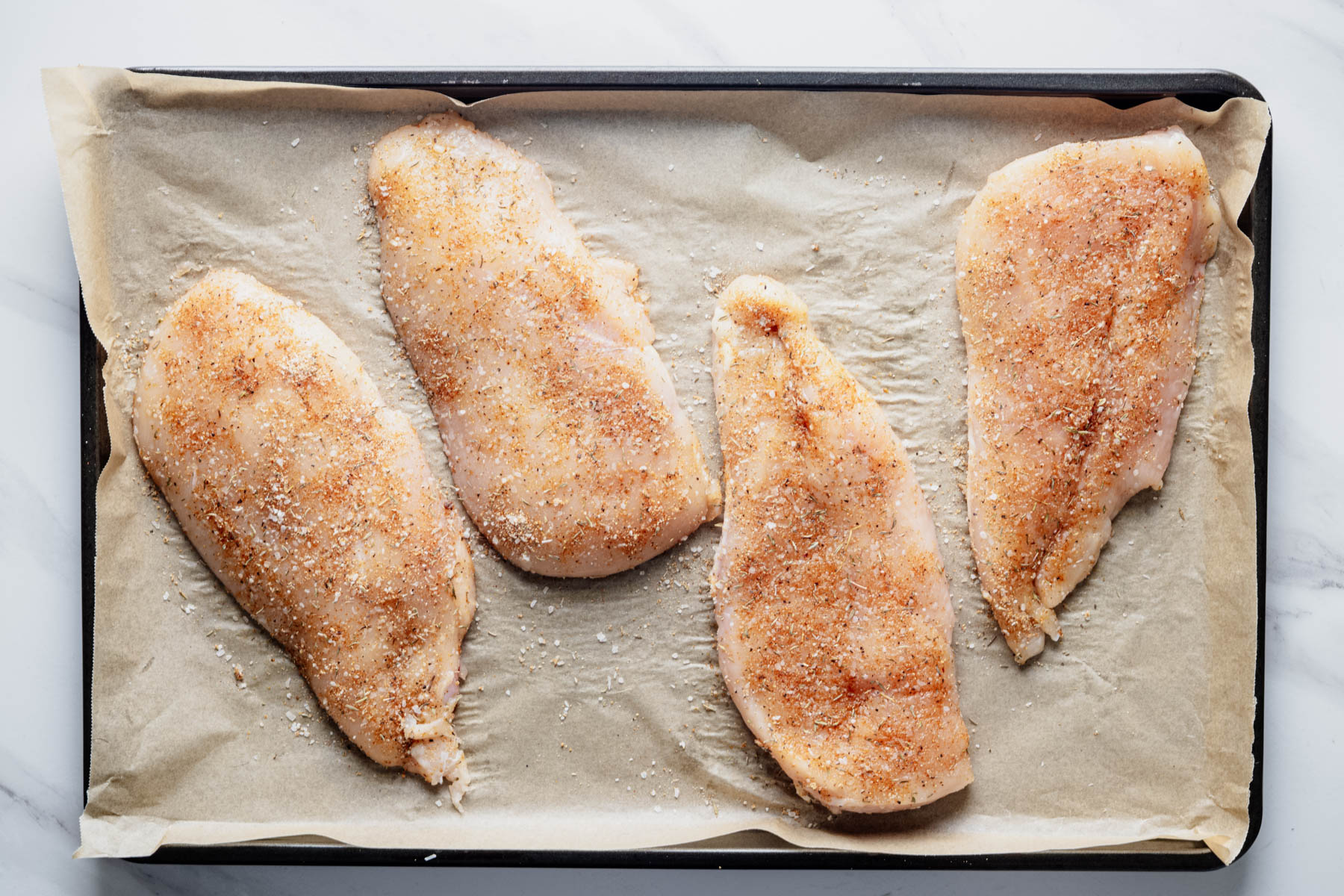 Raw chicken cutlets seasoned with a garlic herb dry brine on a parchment-lined baking sheet before baking.