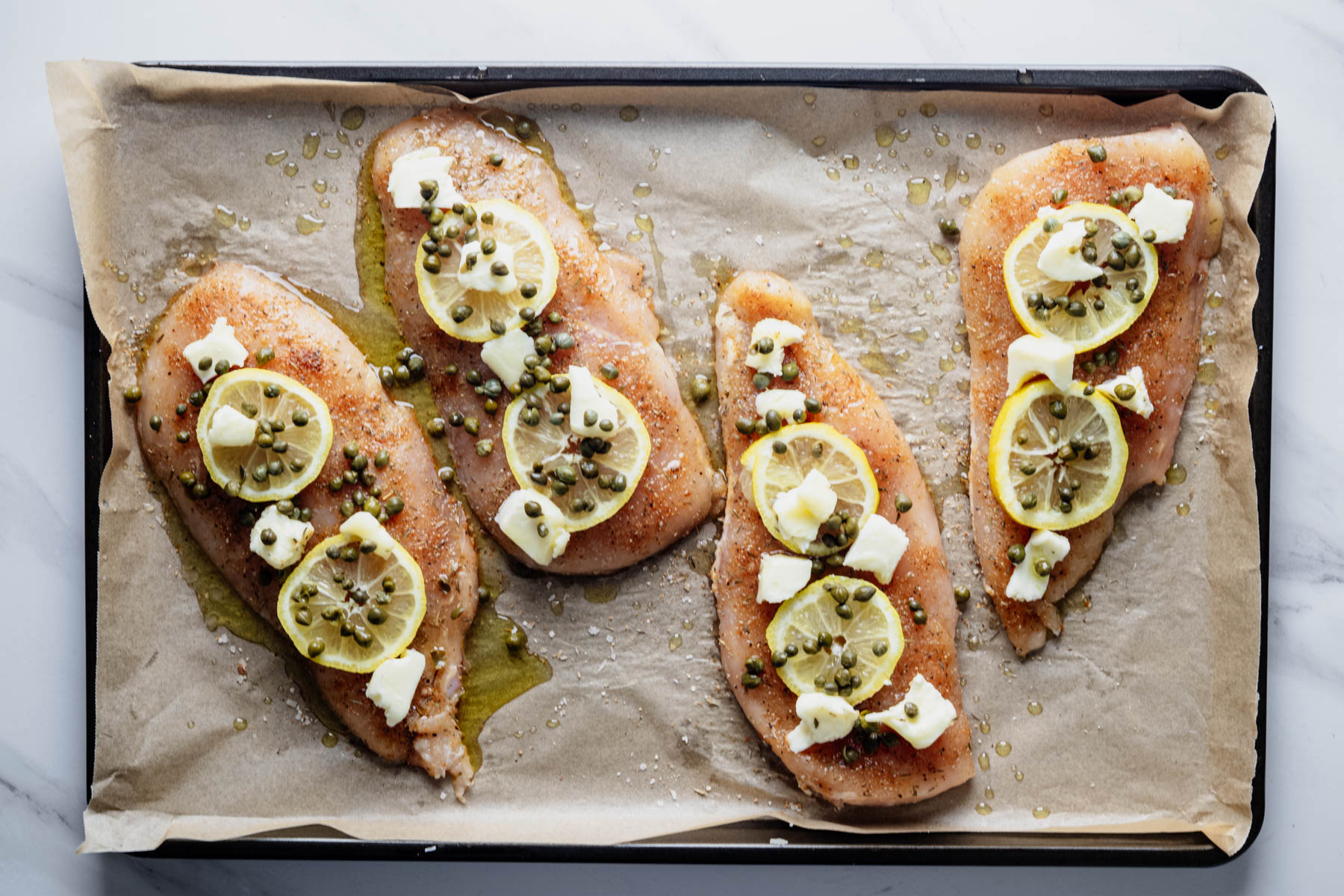 Chicken cutlets on a parchment-lined baking sheet topped with lemon slices, capers, butter pieces, and olive oil before baking for chicken piccata.