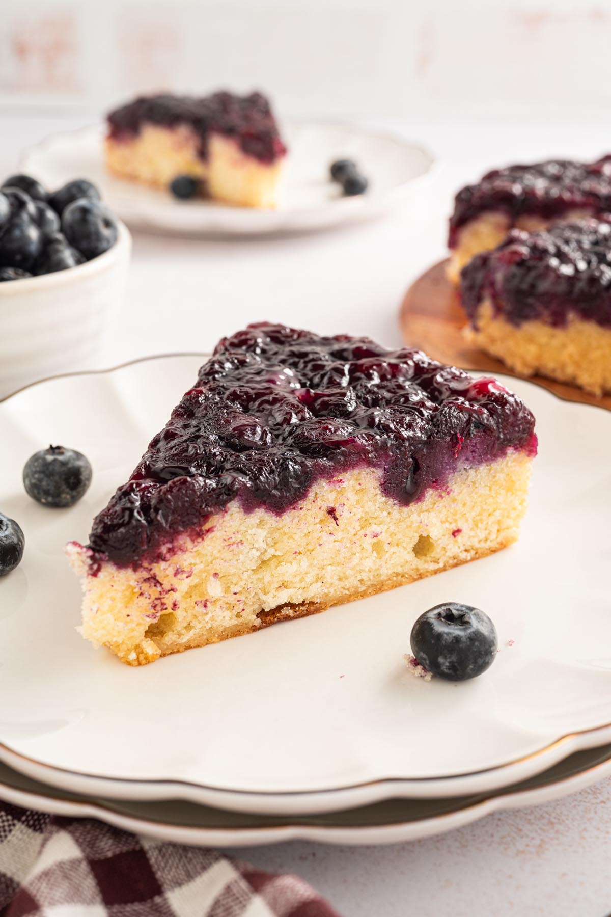 Slice of blueberry upside-down cake on a white scalloped plate with fresh blueberries and additional slices blurred in the background.