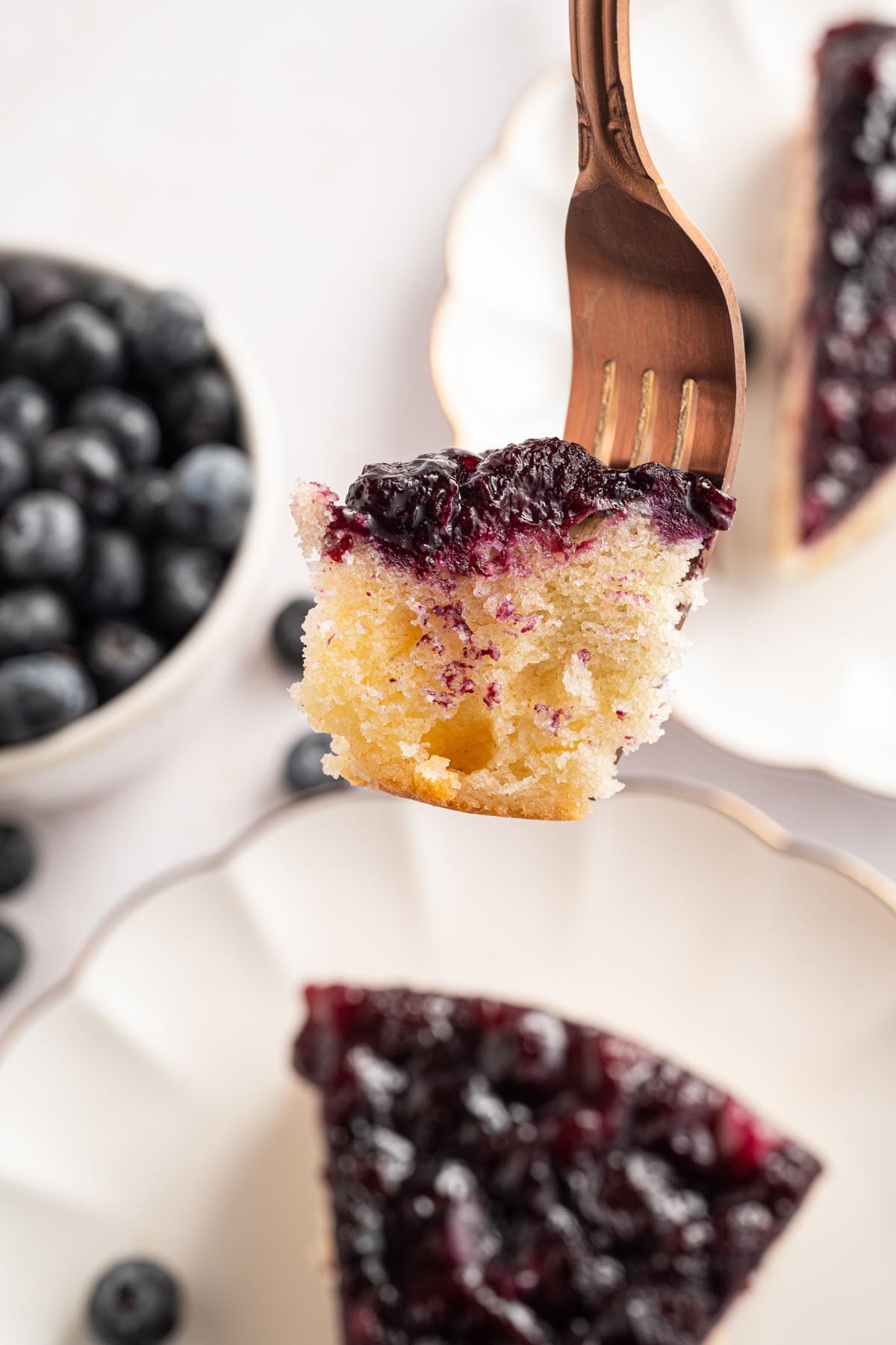 Bite of blueberry upside-down cake held on a copper fork with fresh blueberries and a slice of cake in the background.
