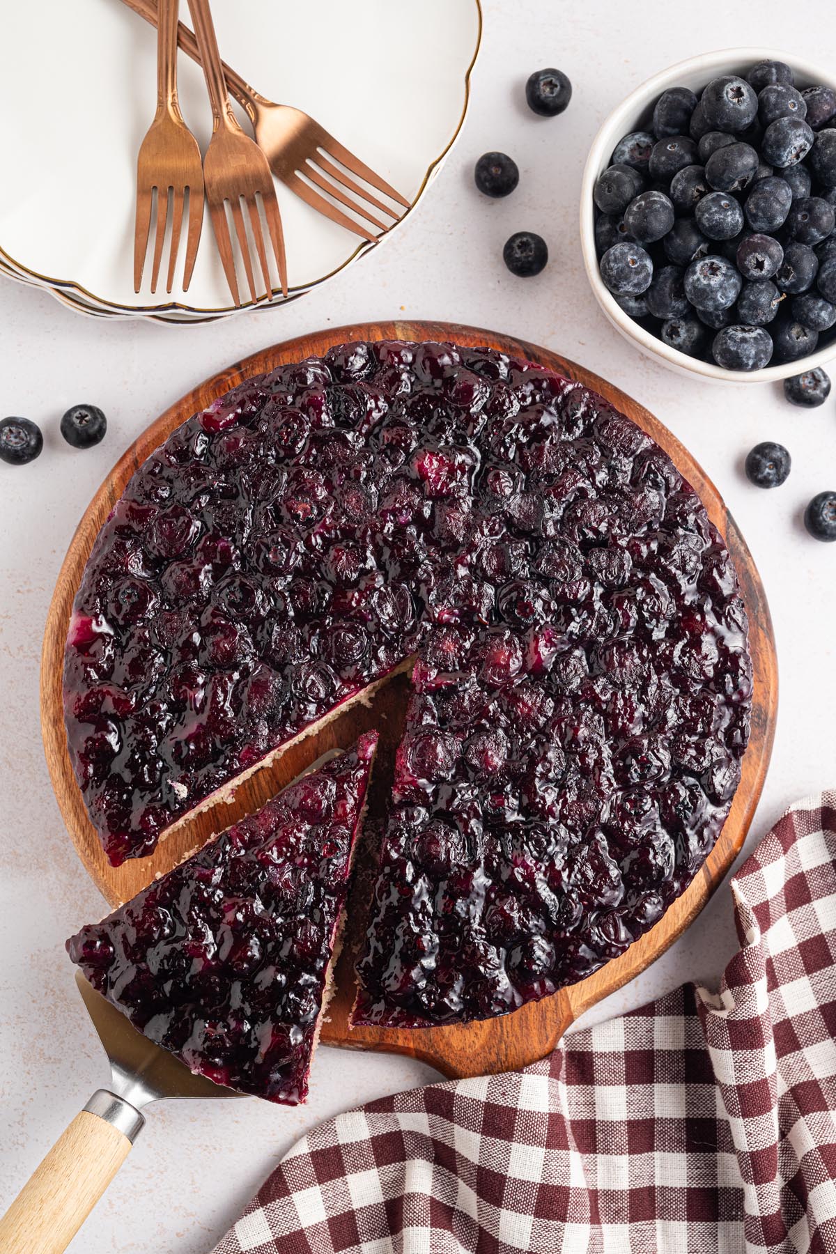 Overhead view of a blueberry upside-down cake on a wooden serving board with fresh blueberries and copper dessert forks nearby.