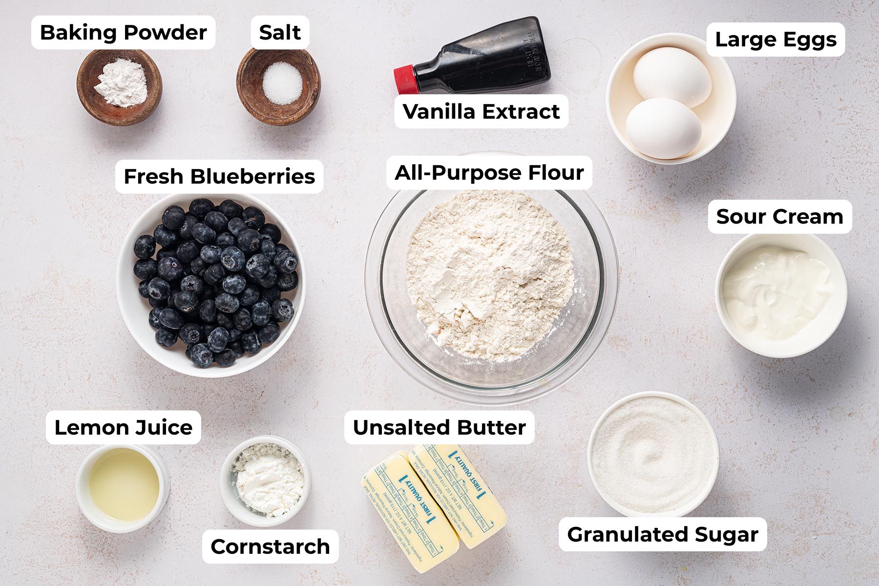 Labeled ingredients for blueberry upside-down cake including fresh blueberries, flour, sugar, butter, eggs, sour cream, lemon juice, cornstarch, vanilla extract, baking powder, and salt arranged on a light surface.