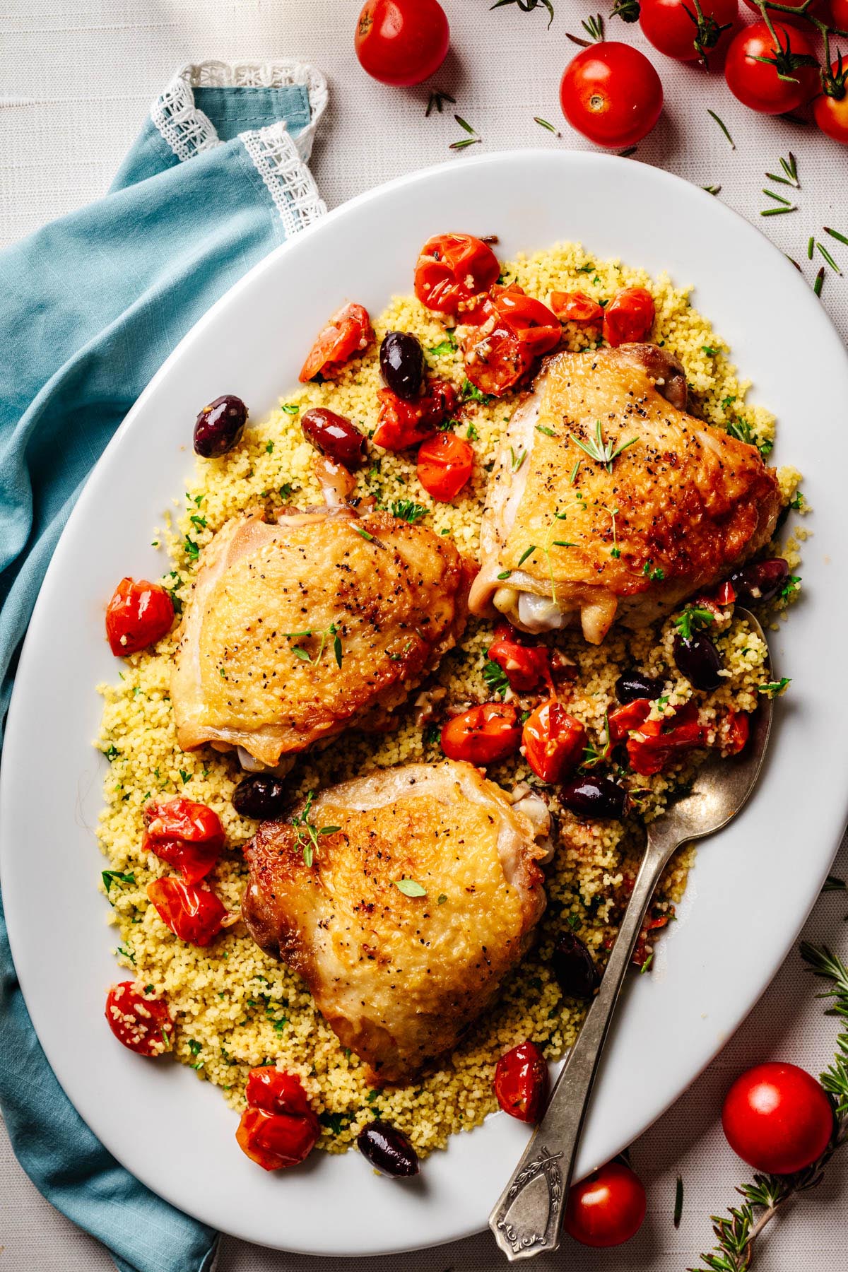 Roasted chicken thighs served on couscous with blistered cherry tomatoes, olives, and fresh herbs on a white platter.