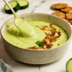 Creamy cucumber gazpacho in a beige bowl with a spoon lifting the soup, topped with crunchy crackers, olive oil, and basil, with cucumber slices and crackers in the background.