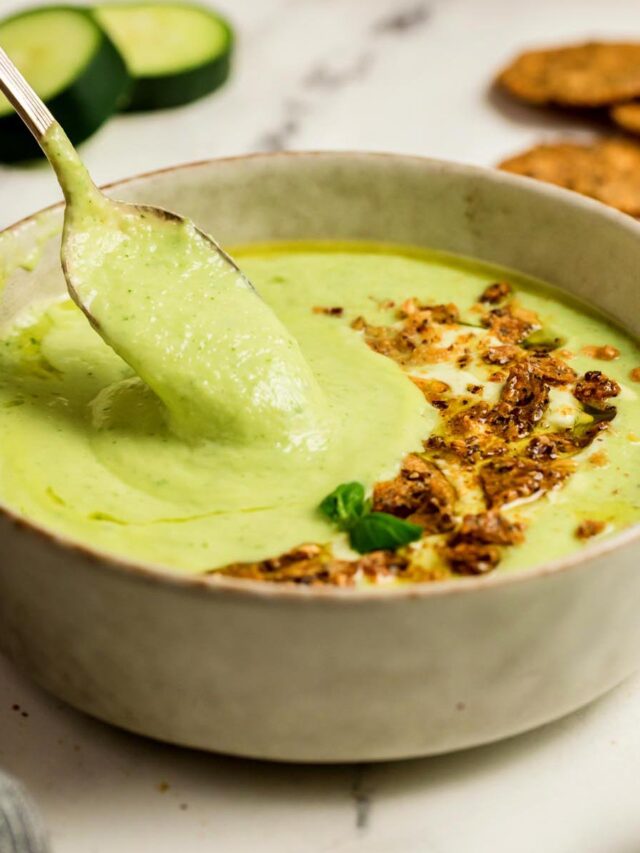 Creamy cucumber gazpacho in a beige bowl with a spoon lifting the soup, topped with crunchy crackers, olive oil, and basil, with cucumber slices and crackers in the background.
