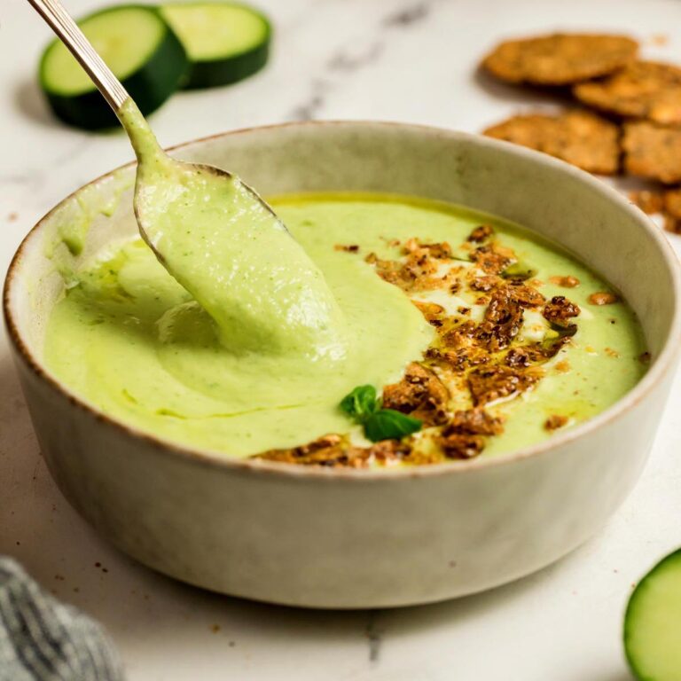Creamy cucumber gazpacho in a beige bowl with a spoon lifting the soup, topped with crunchy crackers, olive oil, and basil, with cucumber slices and crackers in the background.