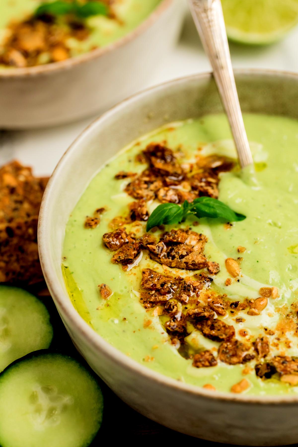Close-up of creamy cucumber gazpacho topped with crunchy crackers, olive oil, and basil in a beige bowl with a spoon dipping into the soup.