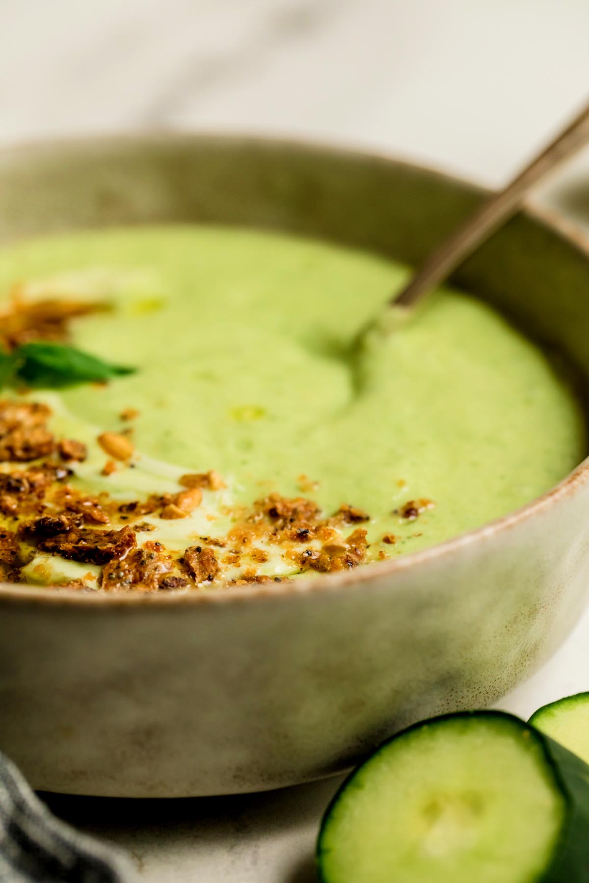 Close-up side view of creamy cucumber gazpacho in a beige bowl topped with crunchy crackers and olive oil with fresh cucumber slices in the foreground.
