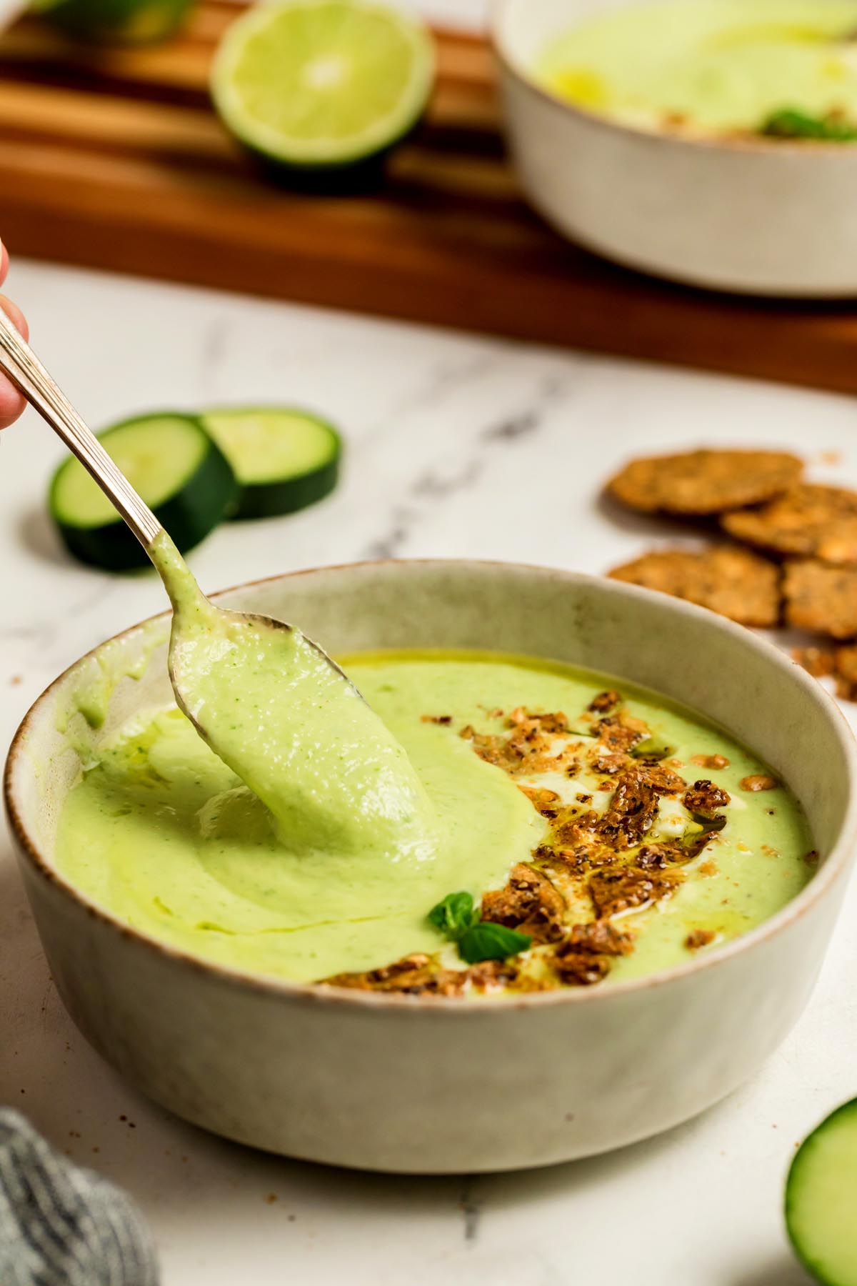Soup in a beige bowl with a spoon lifting the soup, topped with crunchy crackers, olive oil, and basil, with cucumber slices and crackers in the background.