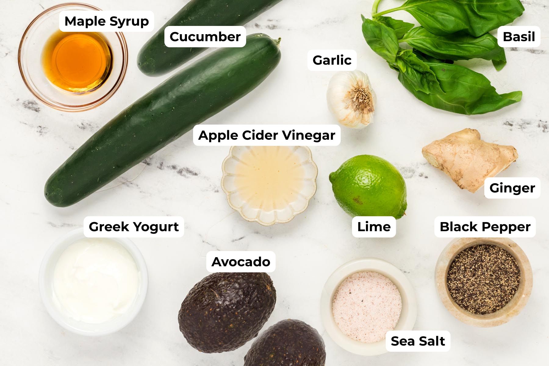 Overhead view of ingredients for cucumber gazpacho, including cucumbers, avocado, Greek yogurt, basil, garlic, ginger, lime, apple cider vinegar, maple syrup, sea salt, and black pepper on a marble surface.