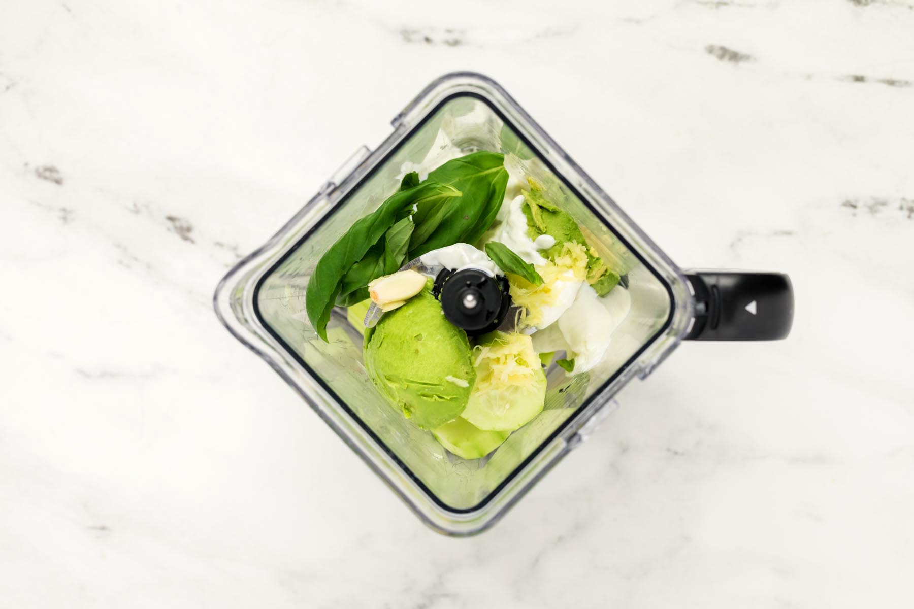 Overhead view of blender filled with ingredients including cucumber, avocado, basil, Greek yogurt, garlic, and ginger before blending.