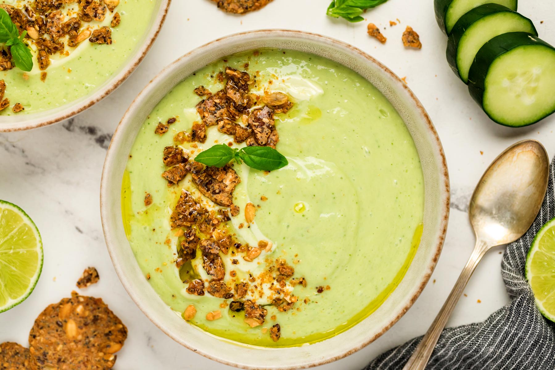 Creamy gazpacho topped with yogurt, olive oil, basil, and crunchy crackers in a beige bowl with cucumber slices and lime on the side.