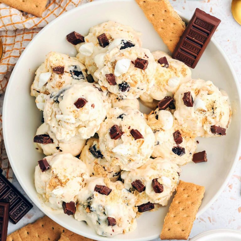 Overhead view of homemade s&rsquo;mores ice cream scoops topped with chocolate chunks, mini marshmallows, and graham cracker crumbs in a white bowl surrounded by graham crackers and chocolate bars.