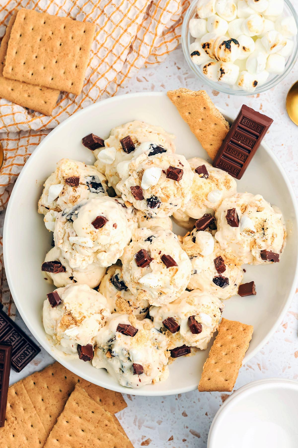 Scoops of homemade s’mores ice cream topped with mini marshmallows, chopped chocolate, and graham cracker crumbs in a white bowl surrounded by graham crackers and chocolate bars.