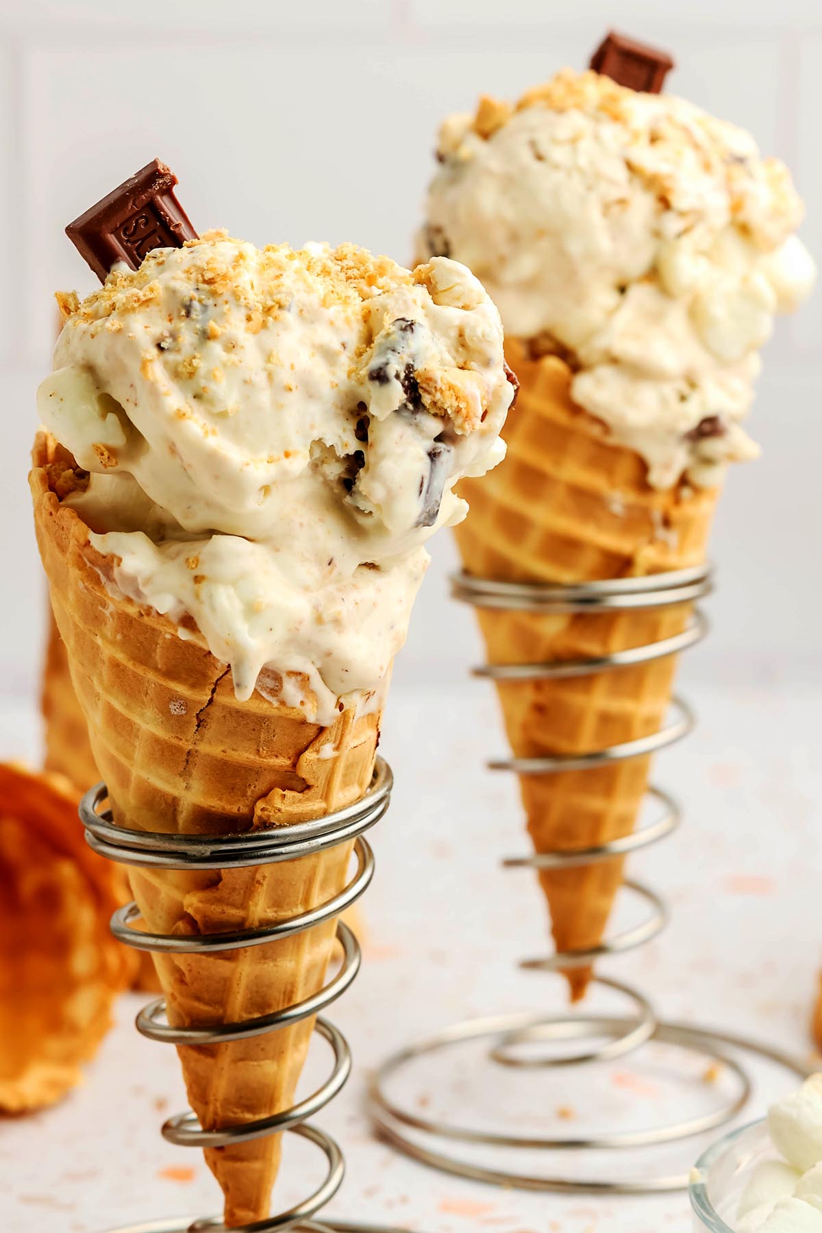 Two waffle cones filled with homemade s’mores ice cream topped with chocolate chunks and graham cracker crumbs in metal cone holders.