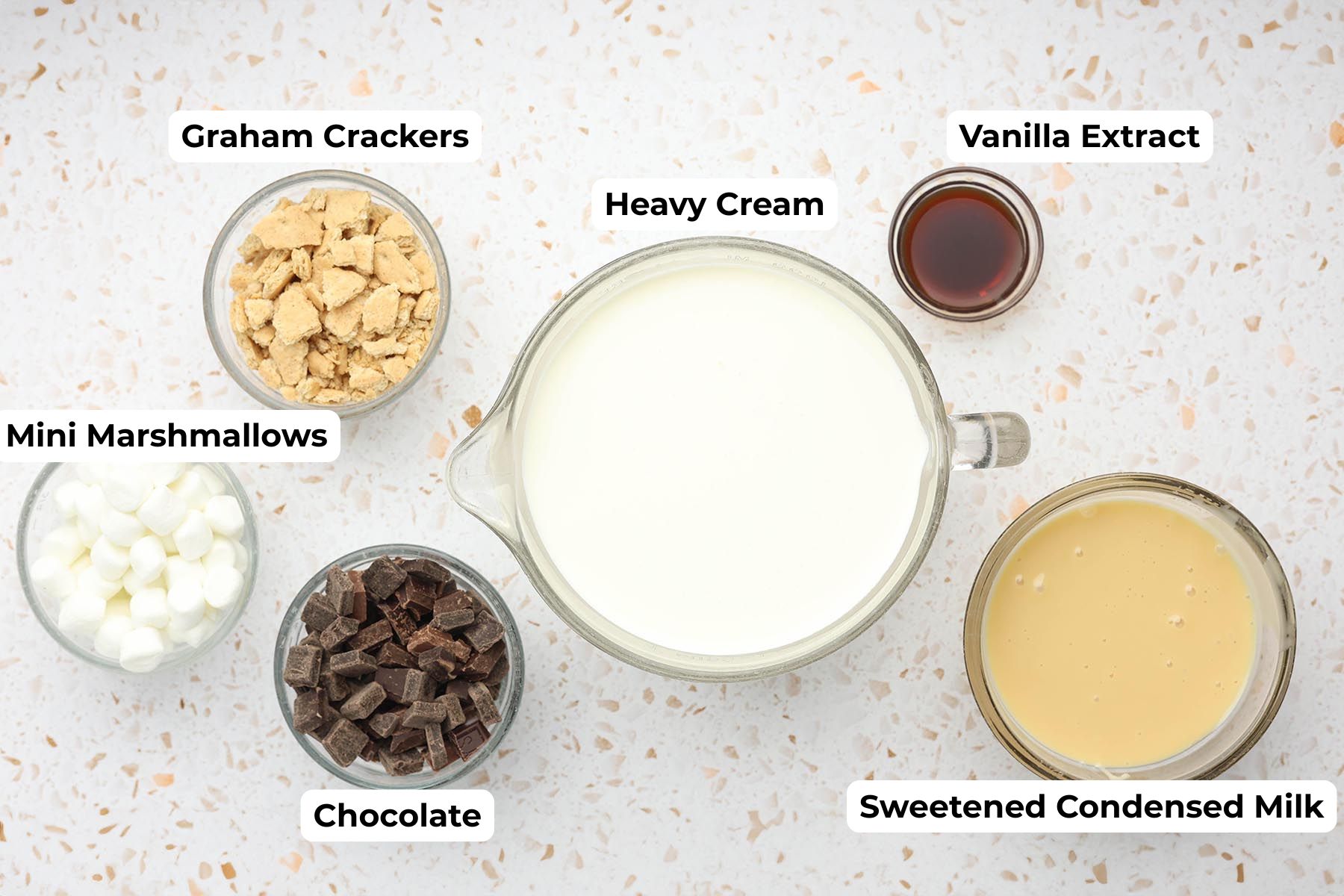 Ingredients for homemade s’mores ice cream including heavy cream, sweetened condensed milk, vanilla extract, chopped chocolate, mini marshmallows, and graham crackers arranged on a light countertop.