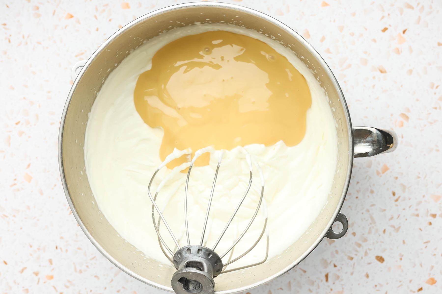 Sweetened condensed milk added to whipped cream in a stand mixer bowl.