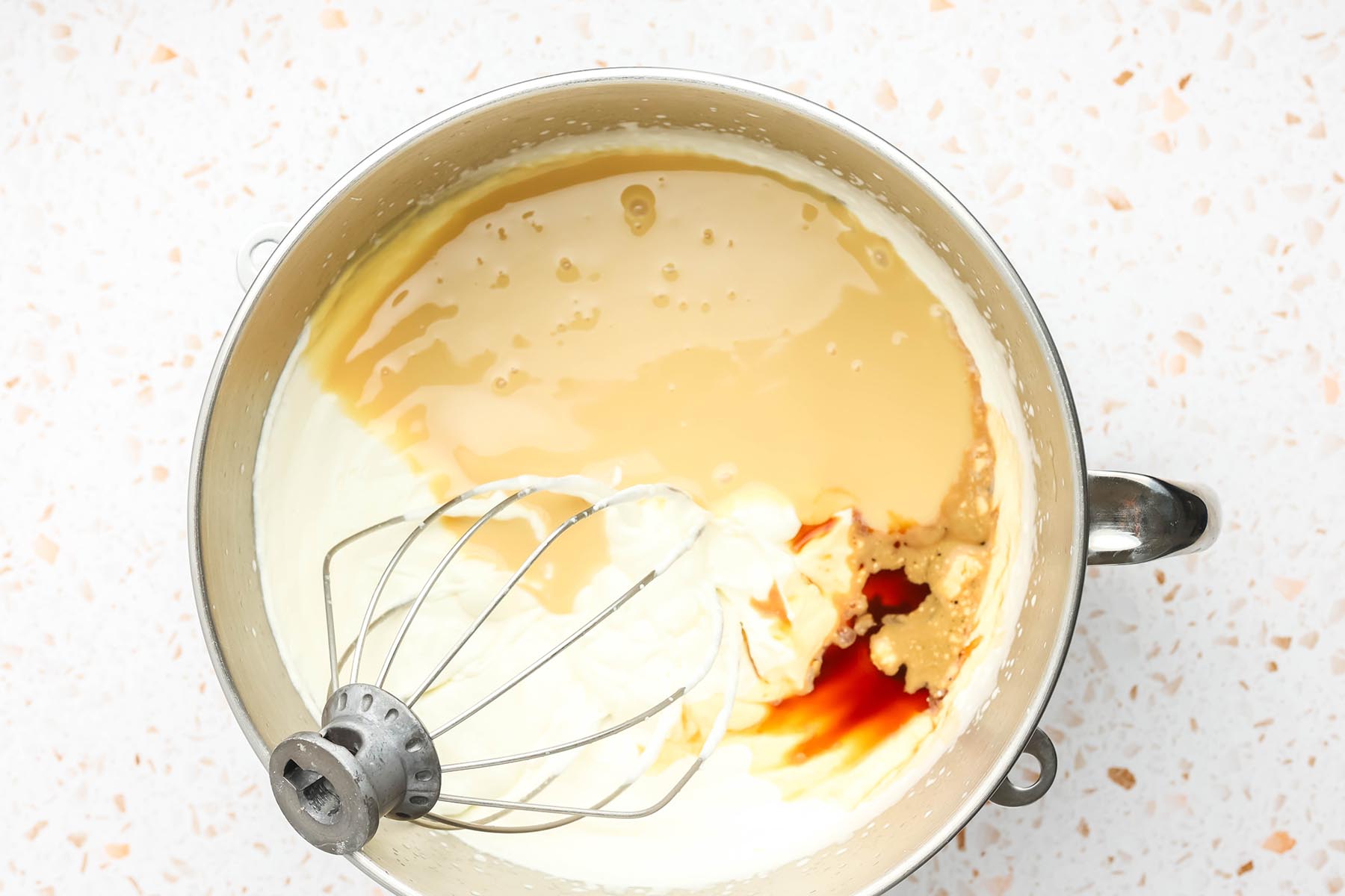 Vanilla extract added to whipped cream and sweetened condensed milk in a stand mixer bowl.