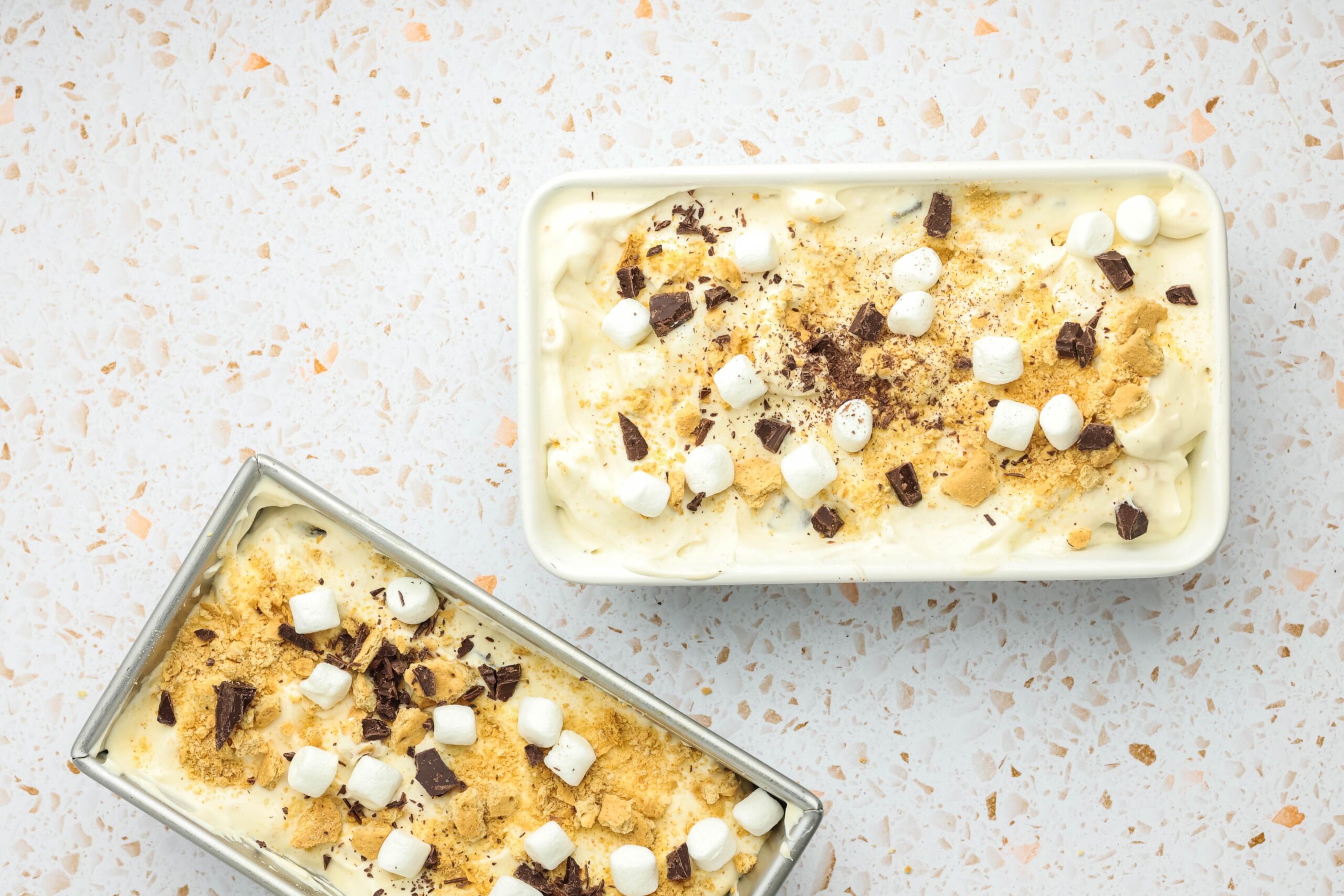 Two loaf pans filled with homemade s’mores ice cream topped with mini marshmallows, chopped chocolate, and crushed graham crackers on a light speckled surface.