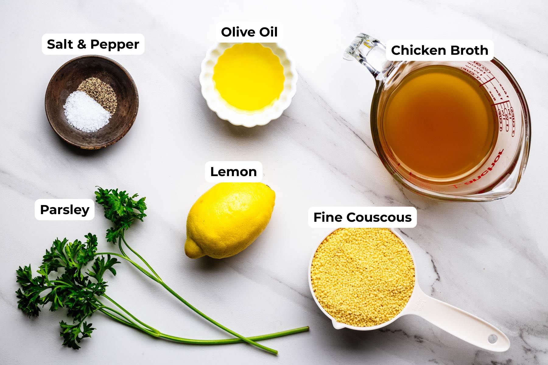 Flat lay of ingredients for lemon couscous including fine couscous, chicken broth, olive oil, fresh parsley, lemon, salt, and black pepper on a light marble surface with labeled ingredient callouts.