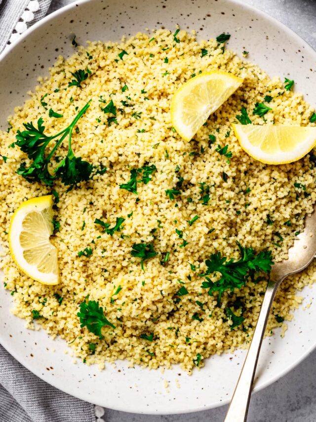 Overhead view of fluffy lemon couscous garnished with fresh parsley and lemon wedges in a large speckled serving bowl with a vintage serving spoon on a light gray surface.