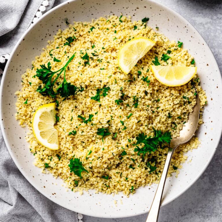 Overhead view of fluffy lemon couscous garnished with fresh parsley and lemon wedges in a large speckled serving bowl with a vintage serving spoon on a light gray surface.