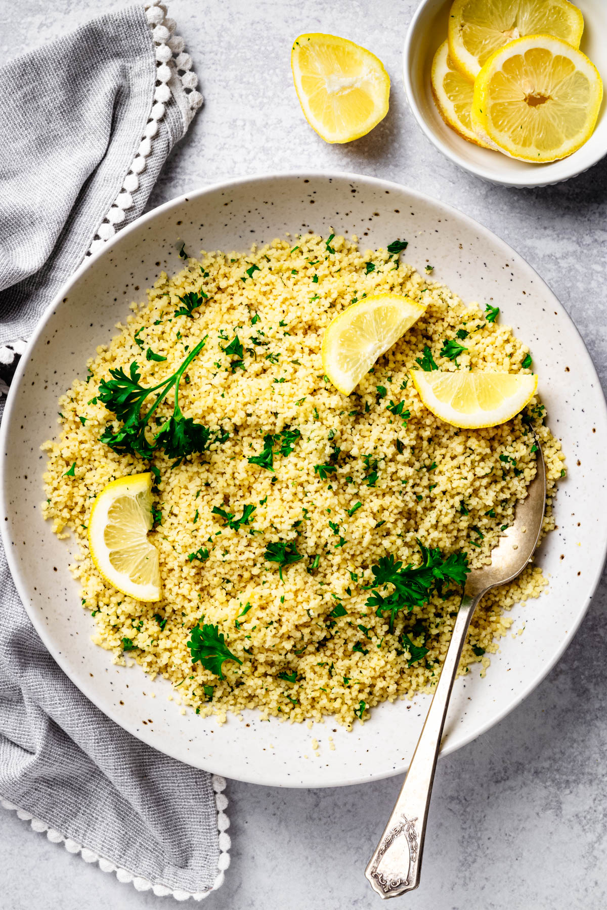 Overhead view of lemon couscous with parsley, lemon wedges, and a serving spoon in a large speckled bowl on a light gray background.