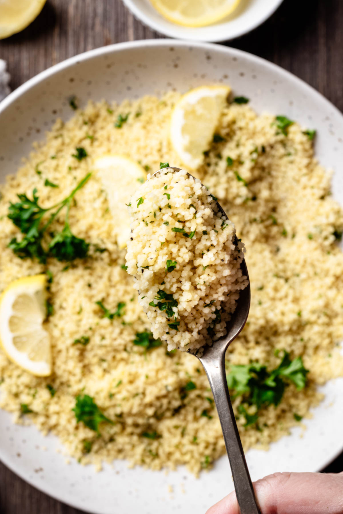 Spoonful of fluffy lemon couscous held above a serving bowl with parsley and lemon wedges on a dark wooden surface.