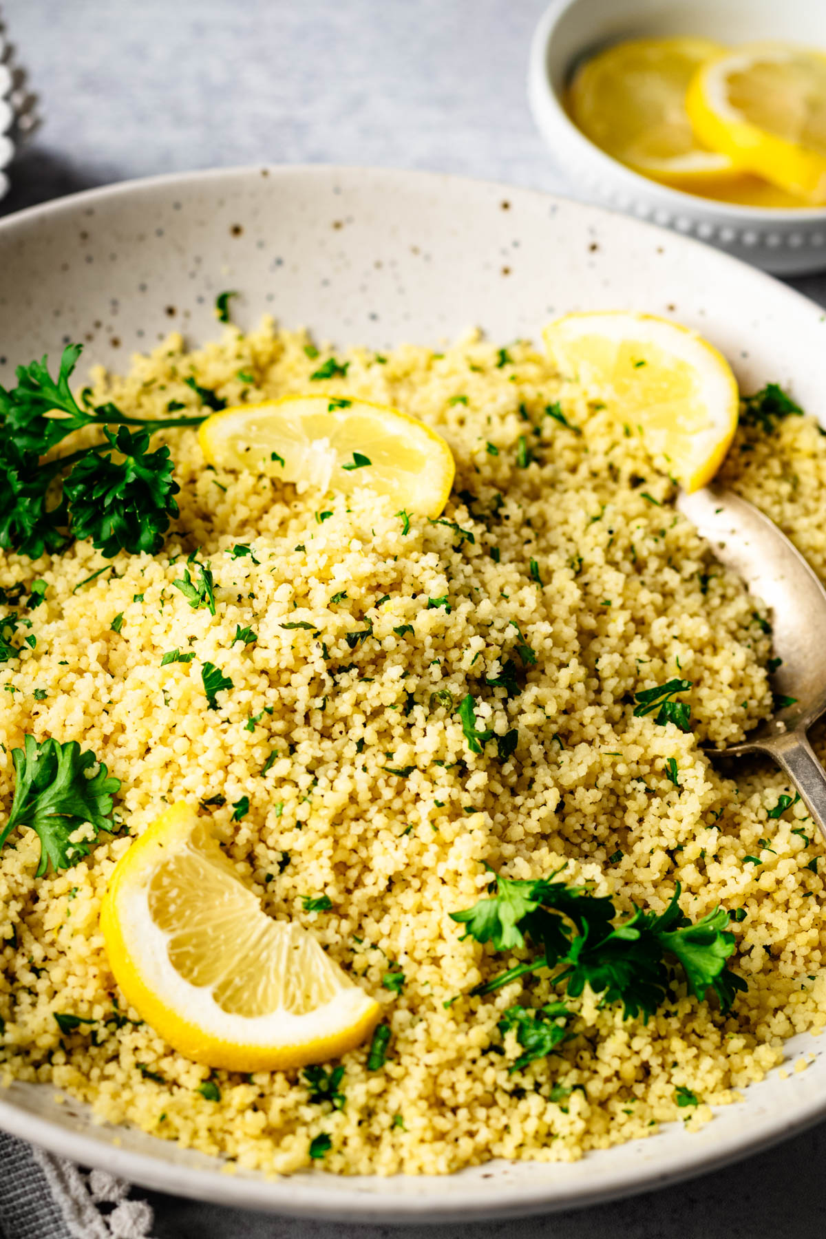 Close side view of fluffy lemon couscous garnished with fresh parsley and lemon wedges in a speckled serving bowl on a light gray surface.